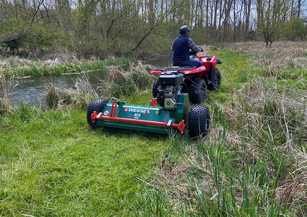 Desmalezadora de arrastre Wessex con motor propio, remolcada por un cuatriciclo, cortando pasto al borde de un arroyo